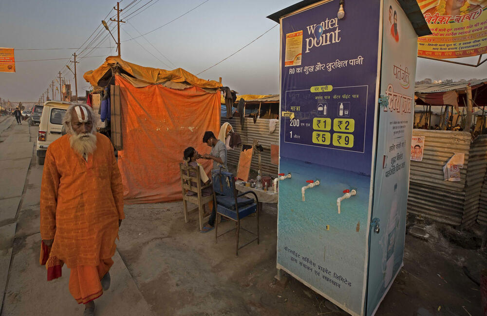 A sadhu walks by a water point at Prayag Kumbh while a roadside barber gives a shave to a customer near a makeshift shelter, 2019. 